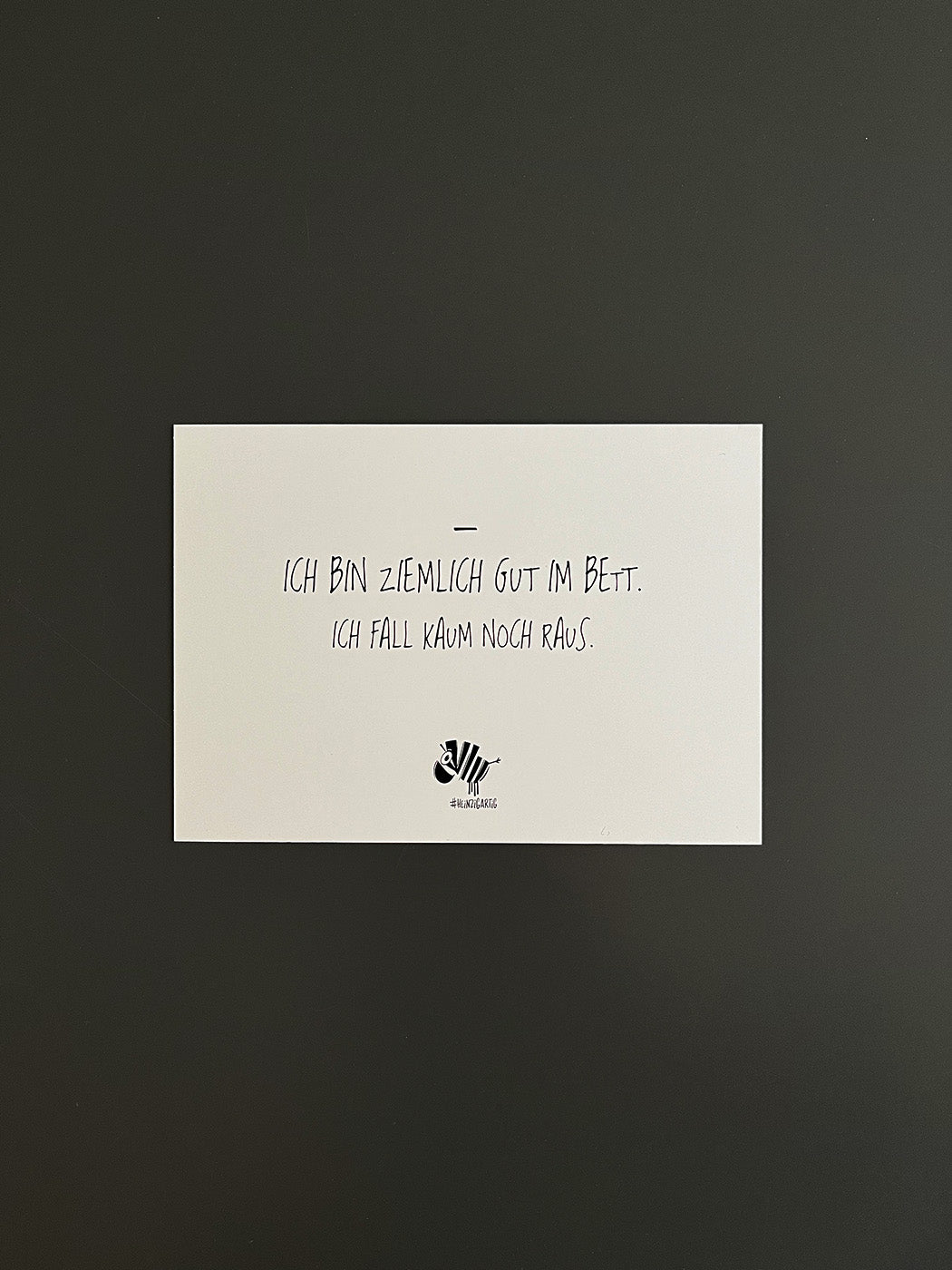 Weiße Postkarte von #heinzigartig mit dem Spruch: "Ich bin ziemlich gut im Bett. Ich fall kaum noch raus."