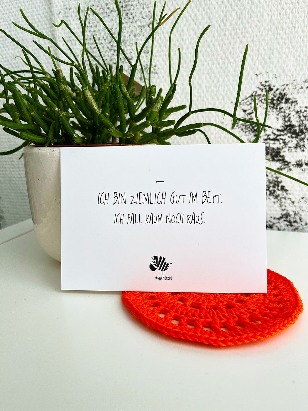 Weiße Postkarte von #heinzigartig mit dem Spruch: "Ich bin ziemlich gut im Bett. Ich fall kaum noch raus."