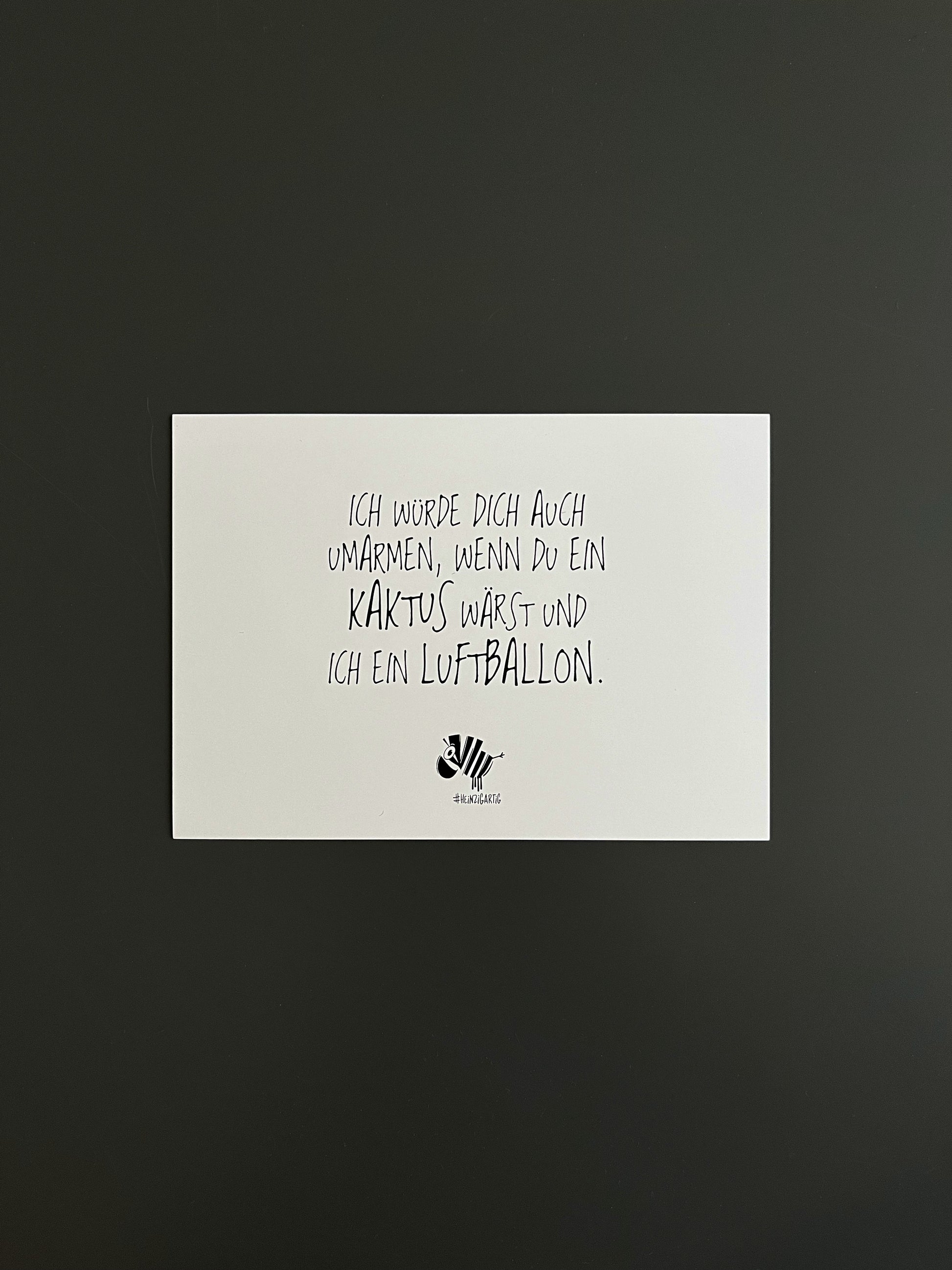 Weiße Postkarte von #heinzigartig mit dem Spruch: "Ich würde dich auch umarmen, wenn du ein Kaktus wärst und ich ein Luftballon."